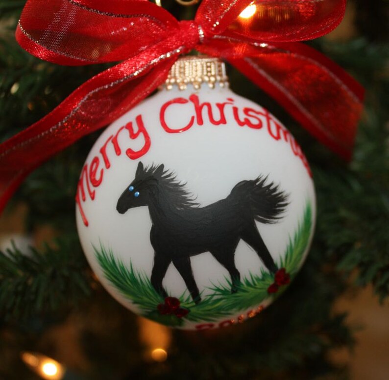 K&ouml;nnte beinhalten: Wei&szlig;er Weihnachtskugel mit einer schwarzen Pferdeillustration und dem Schriftzug "Merry Christmas" in Rot. Der Schmuck hat ein rotes Band und h&auml;ngt an einem Weihnachtsbaum.
