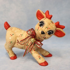May include: A ceramic reindeer figurine with a cream-colored body adorned with red berries and white polka dots. It has red antlers, ears, and hooves, and a red and green ribbon with a gold bell around its neck. The reindeer has large, expressive eyes.