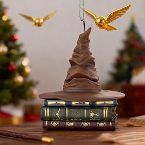 May include: A brown Sorting Hat ornament sits atop a stack of three decorative books. Two golden Snitches are suspended in the air. The books are blue, green, and burgundy with gold accents. A Christmas tree is visible in the background.