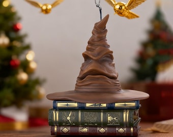 Könnte beinhalten: Ein brauner Sortierhut-Ornament liegt auf einem Stapel von drei dekorativen Büchern. Zwei goldene Schnatze sind aufgehängt. Die Bücher sind blau, grün und weinrot mit goldenen Akzenten. Im Hintergrund ist ein Weihnachtsbaum zu sehen.