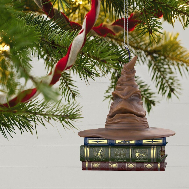 May include: A brown Sorting Hat Christmas ornament, suspended from a silver chain and hanging from a Christmas tree branch. The ornament sits on three stacked books. The books are blue, green, and burgundy. Red and white striped ribbon decorates the tree.