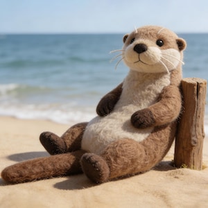 May include: A handcrafted plush otter toy with brown and cream-colored wool. The otter is sitting on a sandy beach, leaning against a wooden post, with the ocean in the background. The toy has a friendly expression.
