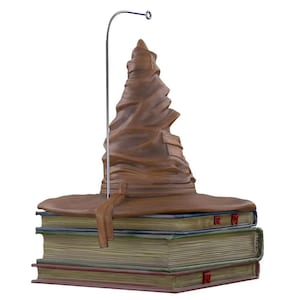 May include: A brown Sorting Hat ornament, with a pointed crown and wide brim, sits on three stacked books. The books have aged pages and red ribbon bookmarks. A silver metal stand extends from the hat. The ornament is a collectible item.