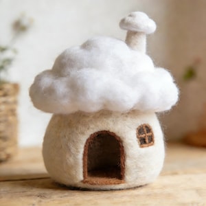 Może przedstawiać: Ręcznie wykonany filcowy domek w kształcie grzyba. Dom jest w kolorze złamanej bieli z puszystym białym dachem w kształcie chmury i małym kominem. Posiada brązowe, łukowate drzwi i małe okno. Całość ma miękki i zachęcający wygląd.