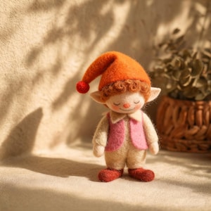 May include: A handmade felt elf doll, featuring an orange hat with a red bobble. The elf wears a pink waistcoat, a beige outfit, and red shoes. The doll has closed eyes and a gentle smile, creating a whimsical appearance.