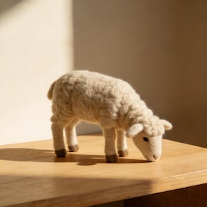 May include: A handcrafted, off-white wool sheep figurine with a textured, curly fleece. The sheep is posed with its head lowered, as if grazing. The figurine has brown hooves and small, dark eyes. It is set on a light brown wooden surface.