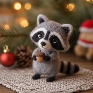 Puede incluir: Figura de mapache de fieltro hecha a mano, gris con marcas negras alrededor de los ojos y una cola a rayas, sosteniendo una pequeña bellota marrón. El mapache está de pie sobre una superficie texturizada de color claro, con un fondo borroso de decoraciones navideñas.