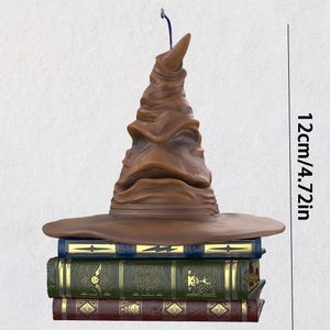 May include: A brown Sorting Hat ornament, 12 cm tall, rests on a stack of three decorative books. The books are blue, green, and burgundy, with gold embossed designs. A blue string is attached to the hat.