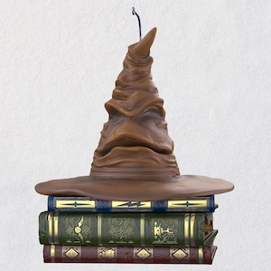 May include: A brown Sorting Hat ornament with a textured finish, perched on a stack of three books. The books are blue, green, and burgundy, with gold detailing. A blue string is attached for hanging.