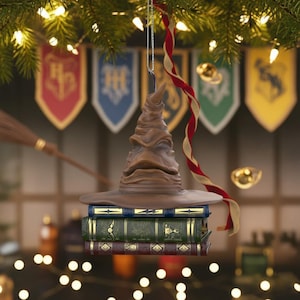 May include: A brown Sorting Hat Christmas tree ornament, suspended by a silver chain, rests on three books. A red and gold ribbon hangs nearby. Banners and a broom are in the background.
