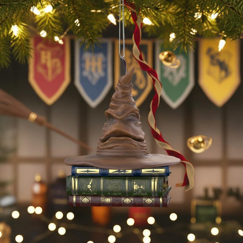 May include: A brown Sorting Hat Christmas tree ornament, suspended by a silver chain, rests on three books. A red and gold ribbon hangs nearby. Banners and a broom are in the background.