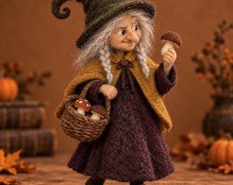Pode incluir: Uma boneca de lã feltrada de uma figura idosa com cabelo grisalho, usando um chapéu pontudo verde-escuro, uma capa amarelo-mostarda e um vestido roxo-escuro. A figura segura um pequeno cogumelo castanho e carrega uma cesta trançada com dois cogumelos.