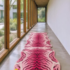May include: Two pink tiger-striped rugs are laid out in a hallway with large windows. The rugs have a light pink background with darker pink and black stripes. The hallway has a concrete floor and wooden window frames. The text "Rug Art USA" is visible.