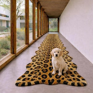 Puede incluir: Una alfombra larga de forma irregular con un estampado de leopardo en tonos marrón, beige y negro. Un cachorro de golden retriever de color claro está sentado en el centro de la alfombra, situada sobre un suelo de hormigón gris claro.