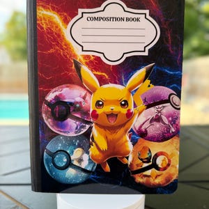 May include: Composition book featuring Pikachu and various Poke Balls against a backdrop of red and blue lightning. The cover has a white label with the words "COMPOSITION BOOK". The notebook is on a white stand.