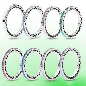 Op de afbeelding: Een verzameling zilveren ringen met hartvormige uitsparingen langs de randen. Sommige ringen hebben een band van gekleurde edelstenen in blauw, roze, groen en paars. De ringen worden gepresenteerd op een groene gradiënt achtergrond.