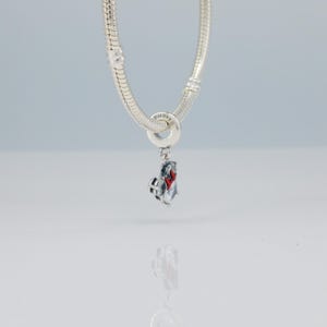 Puede incluir: Una pulsera de plata con un charm colgante. El charm es una peque&ntilde;a figura detallada con un lazo rojo y un logotipo de Disney. La pulsera y el charm est&aacute;n hechos de un metal brillante y reflectante. El fondo es de un color suave y neutro.