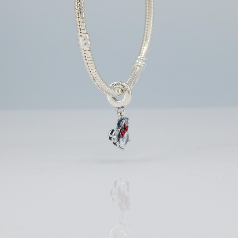 Puede incluir: Una pulsera de plata con un charm colgante. El charm es una peque&ntilde;a figura detallada con un lazo rojo y un logotipo de Disney. La pulsera y el charm est&aacute;n hechos de un metal brillante y reflectante. El fondo es de un color suave y neutro.