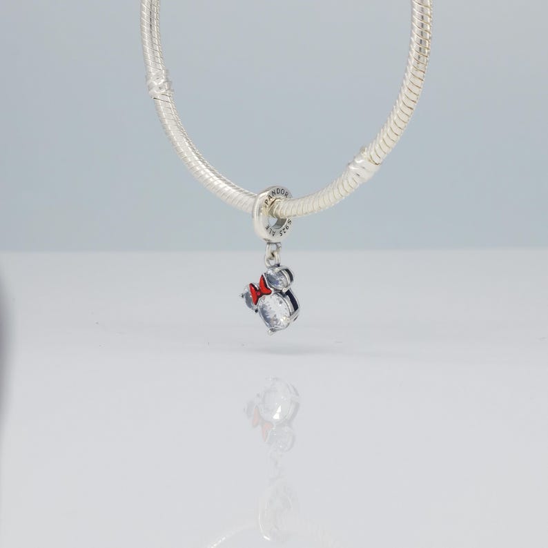 Puede incluir: Un charm de plata con un dise&ntilde;o de Minnie Mouse. El charm presenta un cuerpo de piedra transparente y brillante, un lazo rojo y un lazo plateado para sujetarlo a una pulsera o collar. El charm est&aacute; suspendido de una cadena de plata.