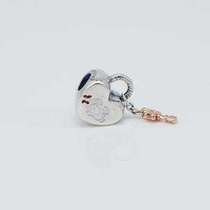 May include: A silver heart-shaped charm with a small rose gold key dangling from it. The charm features a white illustration of a character with red accents. The charm has a loop for attaching to a bracelet or necklace.