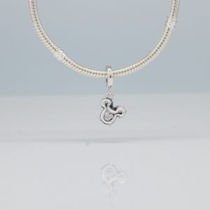 Puede incluir: Un charm de plata con forma de ampersand, colgado de una cadena plateada. El charm, con un acabado pulido, se presenta sobre un fondo claro. Es una pieza de joyer&iacute;a.