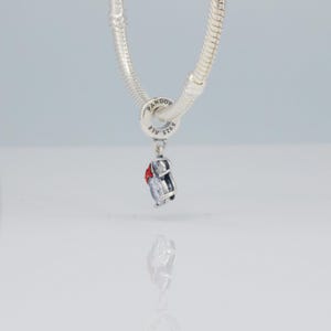 Puede incluir: Un charm de plata con detalles rojos y transparentes, colgando de una cadena de plata. El charm tiene un dise&ntilde;o peque&ntilde;o y detallado. La cadena es de estilo serpiente. El charm es probablemente un accesorio de joyer&iacute;a.