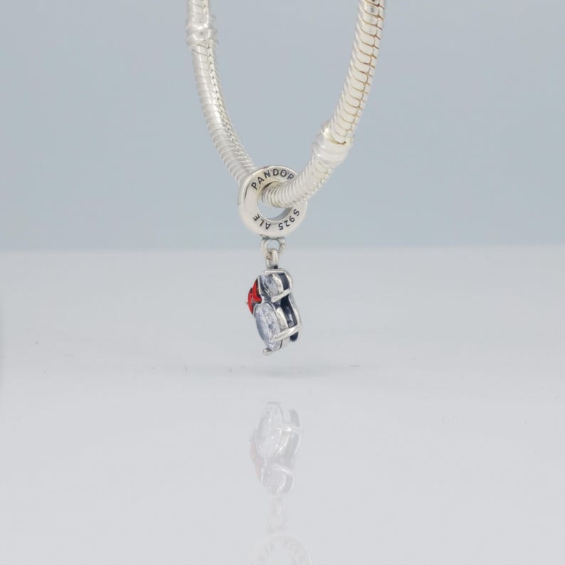 Puede incluir: Un charm de plata con detalles rojos y transparentes, colgando de una cadena de plata. El charm tiene un dise&ntilde;o peque&ntilde;o y detallado. La cadena es de estilo serpiente. El charm es probablemente un accesorio de joyer&iacute;a.