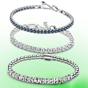 May include: Three silver bracelets. The top bracelet has blue gemstones, the middle one has clear gemstones, and the bottom bracelet has silver beads and lettered cubes that spell "FRIENDS DON'T LIE". A red heart and a green lightning bolt are also included.