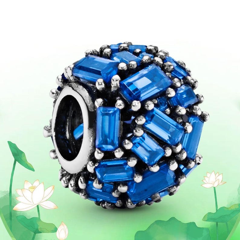 May include: A round charm with a silver-colored base, adorned with numerous rectangular, bright blue gemstones. The gemstones are arranged to cover the entire surface, creating a sparkling effect. Small silver-colored beads surround the blue stones.