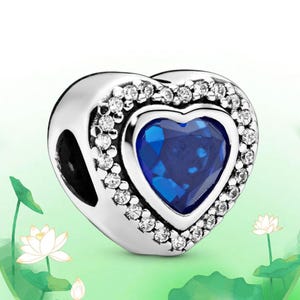 May include: A silver charm with a heart-shaped blue gemstone center, surrounded by a halo of small, clear crystals. The charm has a polished finish and is designed to be added to a bracelet or necklace. The background is a soft green.