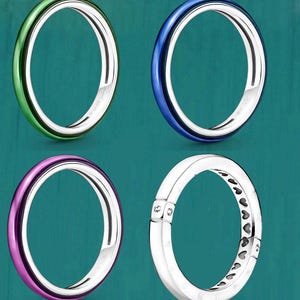 Op de afbeelding: Vier ringen tegen een turquoise achtergrond. Drie ringen hebben een zilveren band met een gekleurde buitenband in groen, blauw en paars. De vierde ring is zilver met een hartpatroon en twee kleine, heldere stenen.