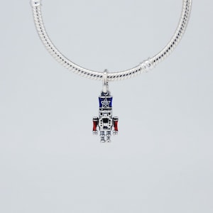 May include: A silver charm in the shape of a toy soldier, with a blue and red uniform and a star on the hat. The charm is suspended from a silver bracelet. The charm is a festive holiday decoration.