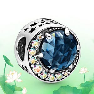 May include: A silver charm with a large, faceted blue gemstone at its center. The charm is encircled by smaller, iridescent gems and features star-shaped cutouts. A decorative piece for bracelets or necklaces.
