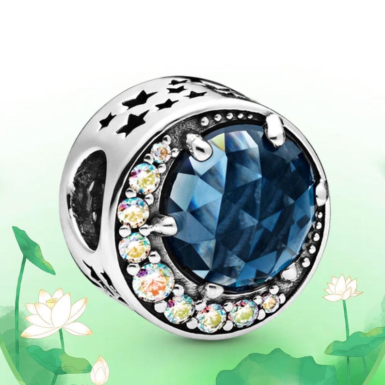 May include: A silver charm with a large, faceted blue gemstone at its center. The charm is encircled by smaller, iridescent gems and features star-shaped cutouts. A decorative piece for bracelets or necklaces.