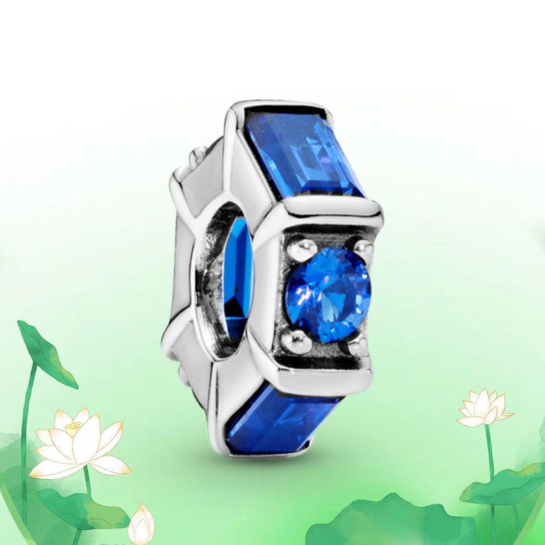 May include: A silver charm with a rectangular blue gemstone and a round blue gemstone. The charm has a geometric design with a polished finish. The gemstones are a vibrant blue color.