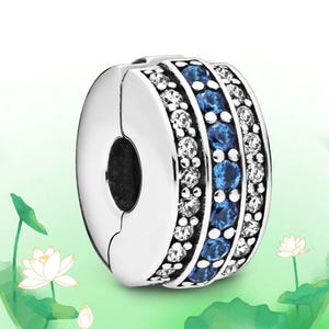 May include: A silver charm with a circular design. The charm features two rows of clear, sparkling stones and a central row of blue stones. The charm is designed to be a clasp, with a split on one side and a black hole in the center.