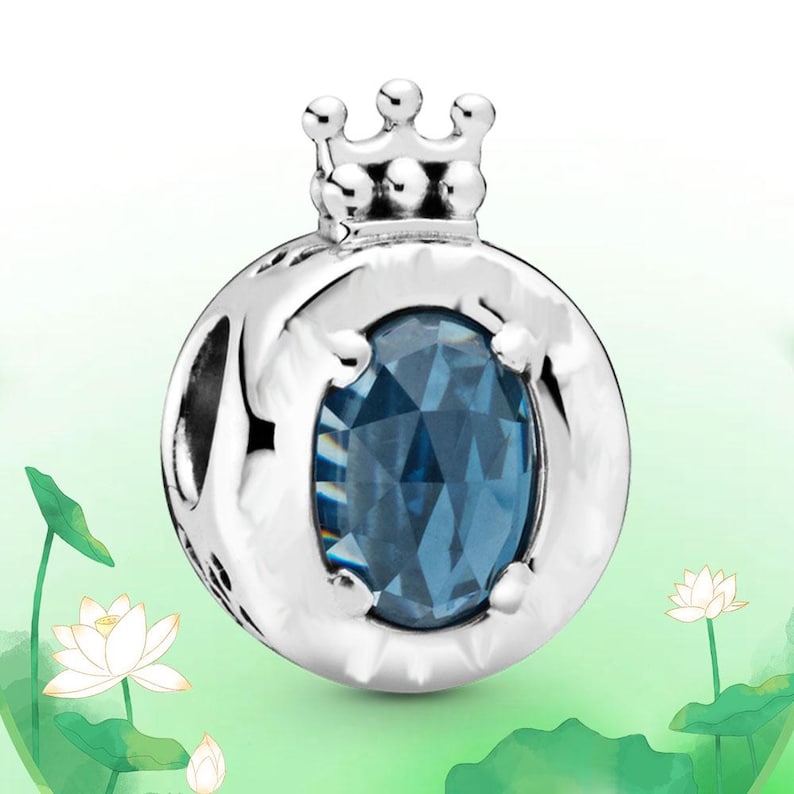 May include: A silver charm with a crown on top and a blue oval-shaped gemstone in the center. The charm has a round shape with cut-out designs and is set against a soft green background with white lotus flowers.