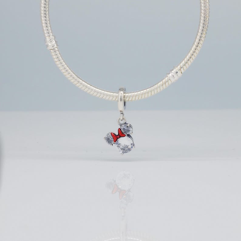 Puede incluir: Un charm de plata con un dise&ntilde;o de Mickey Mouse, con un lazo rojo y detalles de cristal transparente. El charm est&aacute; suspendido de una cadena de plata. Una joya.
