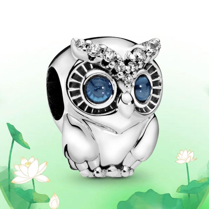 May include: A silver owl charm with blue eyes and sparkling accents on the head. The owl has a detailed design and is set against a soft green background with white lotus flowers.