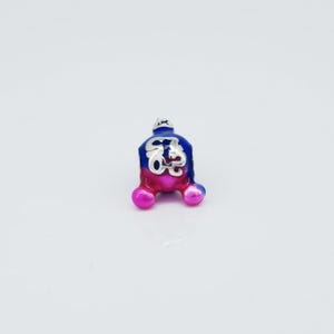 Può includere: Un piccolo ciondolo colorato con un design blu e rosa. Il ciondolo ha una parte superiore color argento e un simbolo decorativo al centro. Il ciondolo ha una forma arrotondata con tre piccoli piedini sferici. Il ciondolo è probabilmente in metallo e smalto.