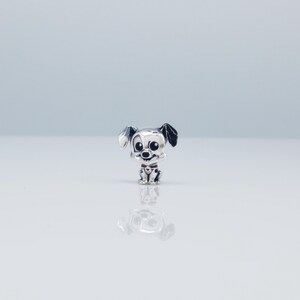 Op de afbeelding: Een kleine, zilverkleurige bedel in de vorm van een Dalmatische puppy. De bedel heeft zwarte vlekken, grote zwarte ogen en een rode halsband. De puppy zit rechtop met opstaande oren.