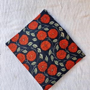 May include: A folded square of fabric with a dark navy background and a repeating pattern of large, orange floral blooms. The flowers have detailed centers and are connected by green leafy vines. The fabric is set against a white textured background.