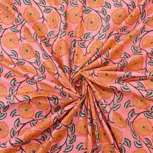May include: A close-up of a pink fabric with a repeating floral pattern. The design features large orange flowers with detailed petals and green leaves connected by dark brown stems. The fabric is folded, creating a textured appearance.