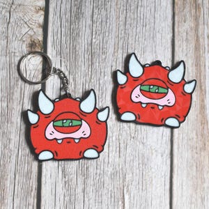 May include: Two red cartoon monster keychains and a matching charm. The monsters have white horns and accents, a single green eye, and a pink mouth. One keychain has a silver ring and chain. The items are on a light wood grain surface.