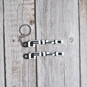 May include: A black and white F150 keychain and emblem set. The keychain has a silver ring and chain. The emblem is a rectangular shape with the text "F150" in a bold, sans-serif font. The background is a light wood grain.