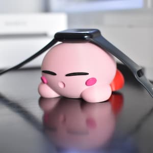 May include: A pink, cartoon-like character with black eyes and pink cheeks, topped with a black smartwatch. The character is lying down on a reflective black surface. The watch band is dark gray.