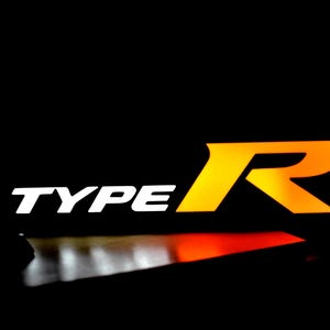 May include: A striking image with the text "TYPE R". The word "TYPE" is in white, while the "R" is a gradient of yellow and orange. The background is a deep black, providing a strong contrast.