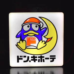 Può includere: Cartello quadrato e illuminato con un pinguino dei cartoni animati con un cappello da Babbo Natale, appollaiato su una luna crescente gialla. Il pinguino è blu con becco e zampe gialle. Il cartello ha testo giapponese in basso e un logo rosso e arancione.