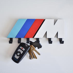 以下が含まれることがあります： BMW Mロゴの形をした壁掛けキーホルダー。キーホルダーは、ライトブルー、ダークブルー、レッドの象徴的なMカラーと白い「M」が特徴です。黒いフックが下に付いており、鍵を保持します。車のキーと他のキーがフックからぶら下がっています。