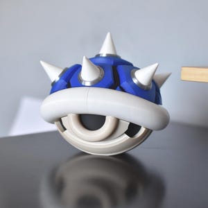 May include: A blue and white spiky Koopa shell from the Mario video game series. The shell has a white rim and spikes, with a blue top. The object is sitting on a reflective black surface.